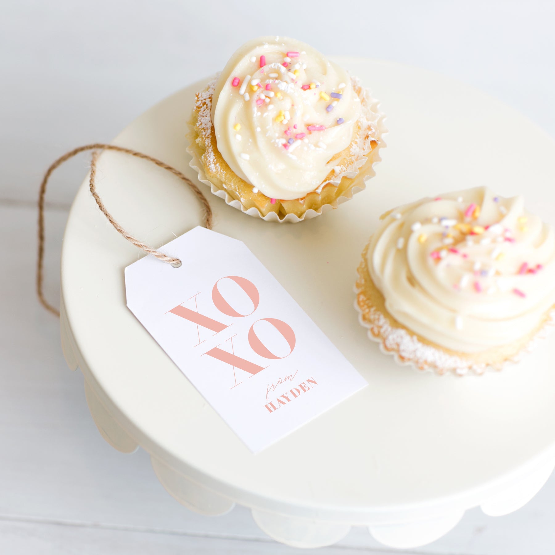 Personalized XOXO gift tag in blush tied to cupcakes with frosting and sprinkles on white cake stand.