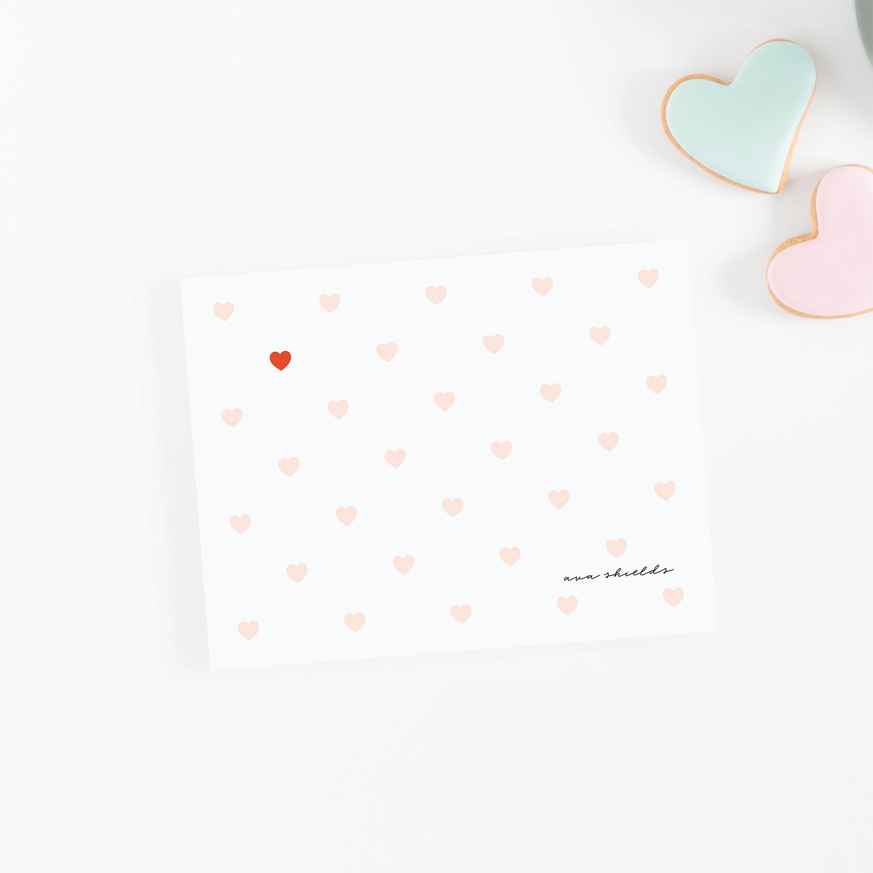 Personalized note card with blush heart pattern and one red accent heart, styled with pastel heart cookies on a white background.