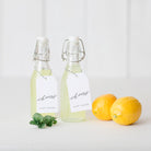 Set of personalized cheers gift tags tied to glass bottles with lemons and mint on white tabletop.