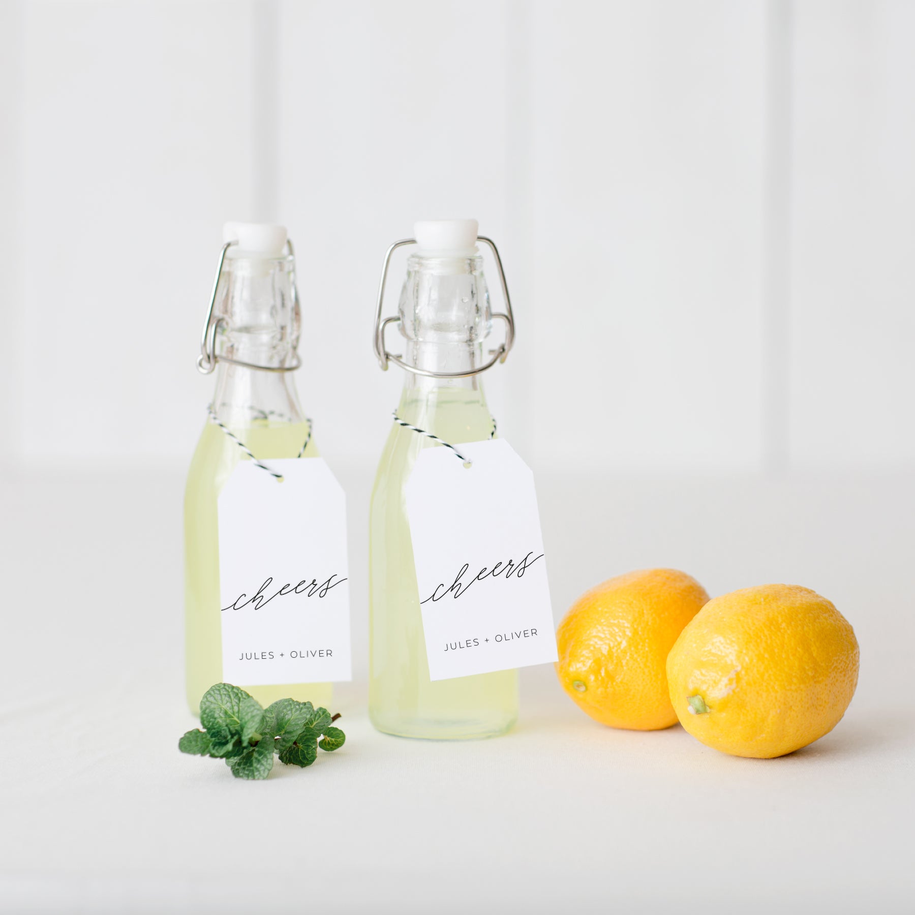 Set of personalized cheers gift tags tied to glass bottles with lemons and mint on white tabletop.