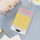 Personalized pencil-inspired notepad with a custom name printed at the top in script font, styled on a light blue desk with gold office accessories, keyboard, and white flowers in the background.