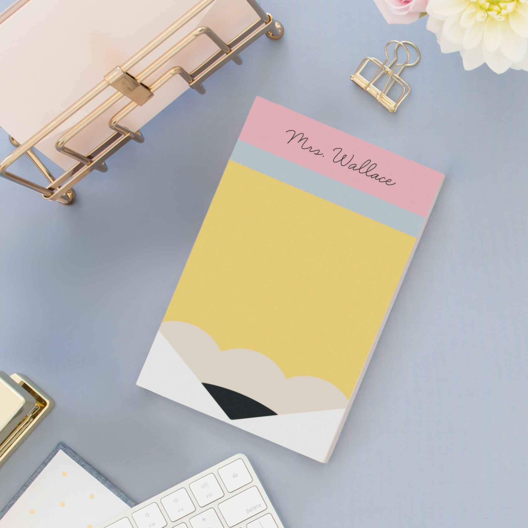 Personalized pencil-inspired notepad with a custom name printed at the top in script font, styled on a light blue desk with gold office accessories, keyboard, and white flowers in the background.