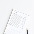 Personalized baby care log notepad with feeding and diaper tracking sections styled on white background with pencil.